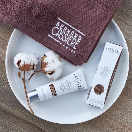 Bernard Cassiere chocolate skincare cream lays on a clean plate alongside two cotton flowers and a Bernard Cassiere towel.