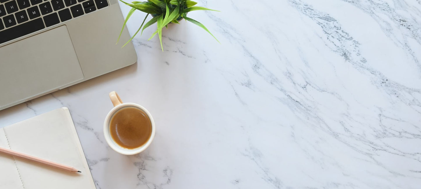 A laptop, notebook, cup of coffee, and a small plant aesthetically placed on a white marble countertop. 