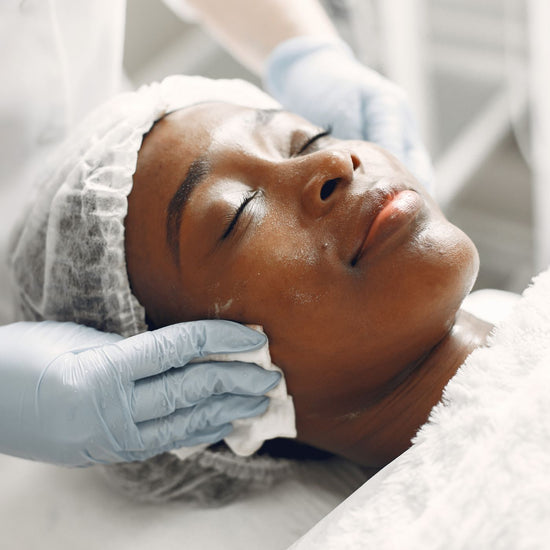 A black woman lays on a table with her eyes closed as she receives a facial treatment from a beauty professional.