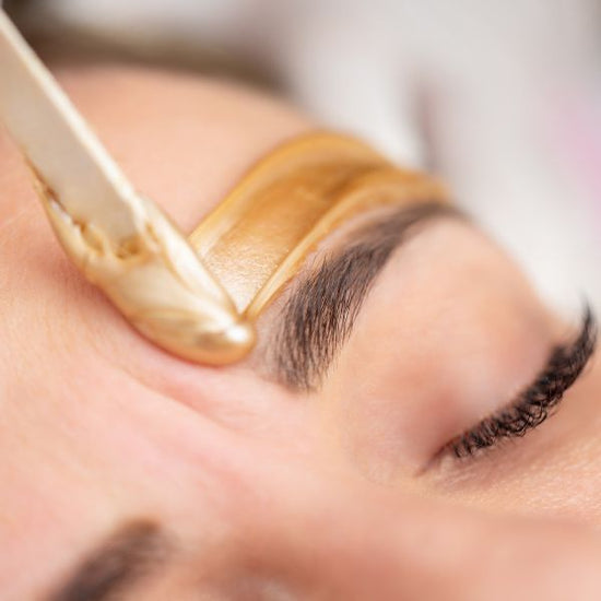 A woman lays on a spa bed with her eyes closed as her certified wax technician applies warm wax above her eyebrow.