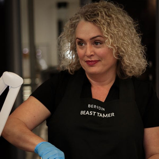 A beauty professional wearing a Berodin apron and gloves uses a wooden stick to mix hot wax.