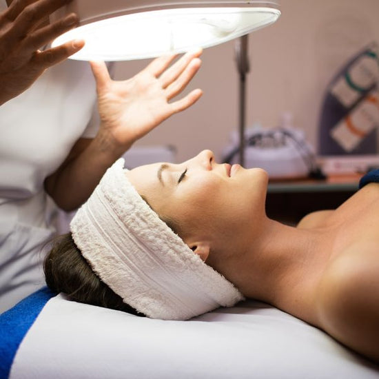 Young women lays on a treatment bed as her facial technician adjusts an overhead light.